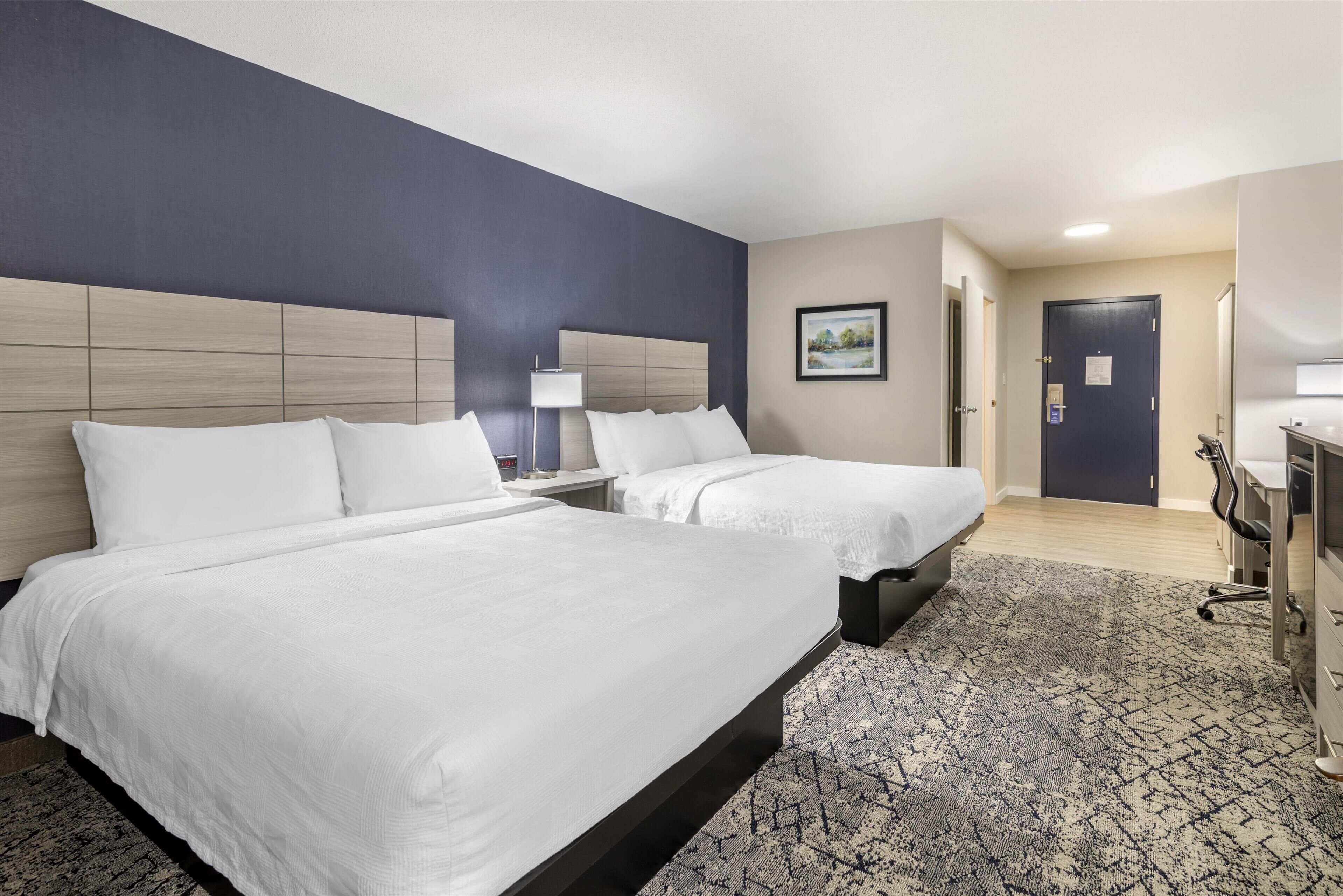 A hotel room with two beds, a desk, a chair, and a door.