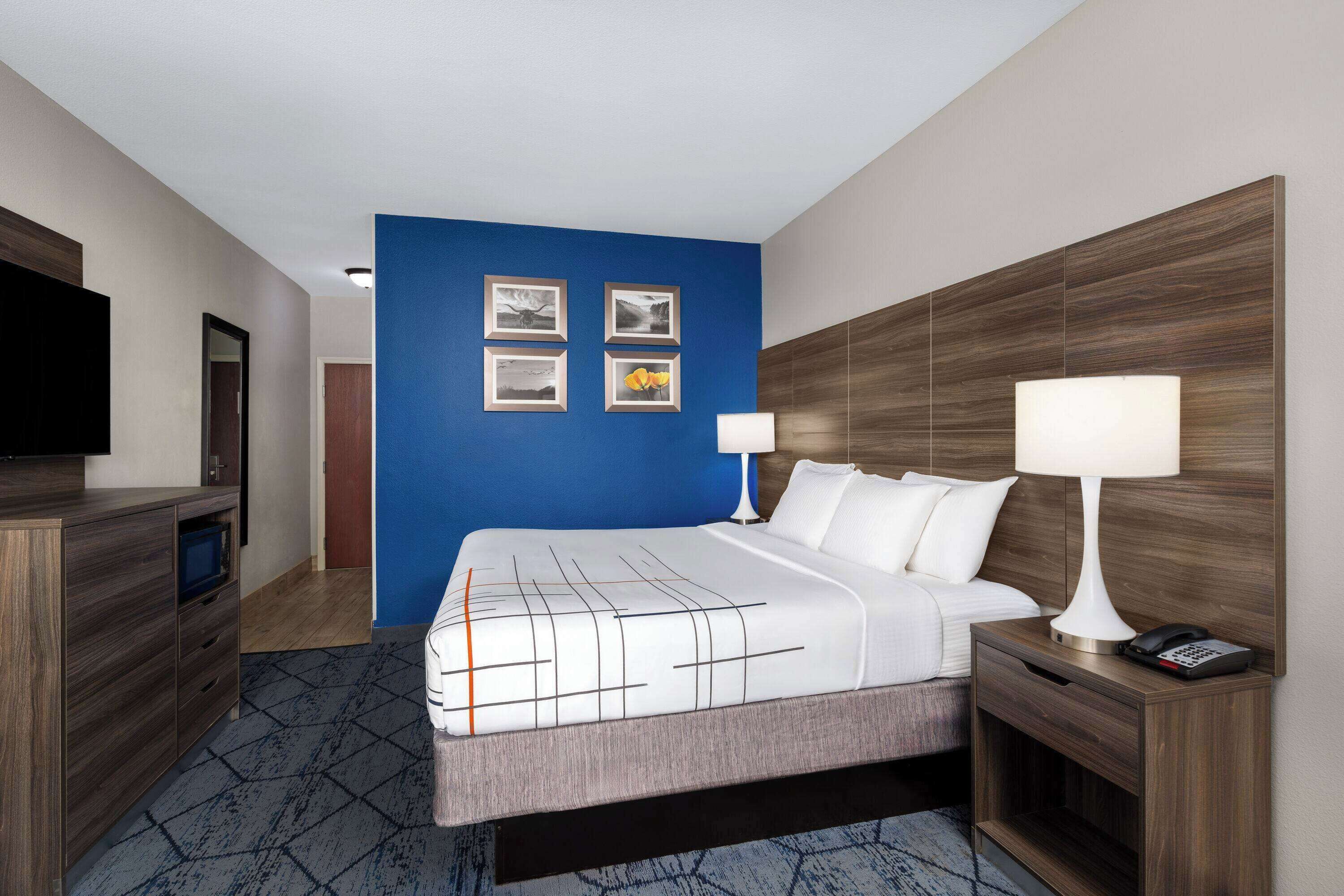 Habitación de hotel con una cama grande, una mesita de noche con lámpara, un televisor sobre un soporte y una pared azul decorada con cuadros.