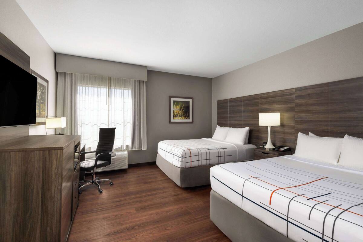 A hotel room with a wooden desk, two beds, a chair, and a large window with curtains.