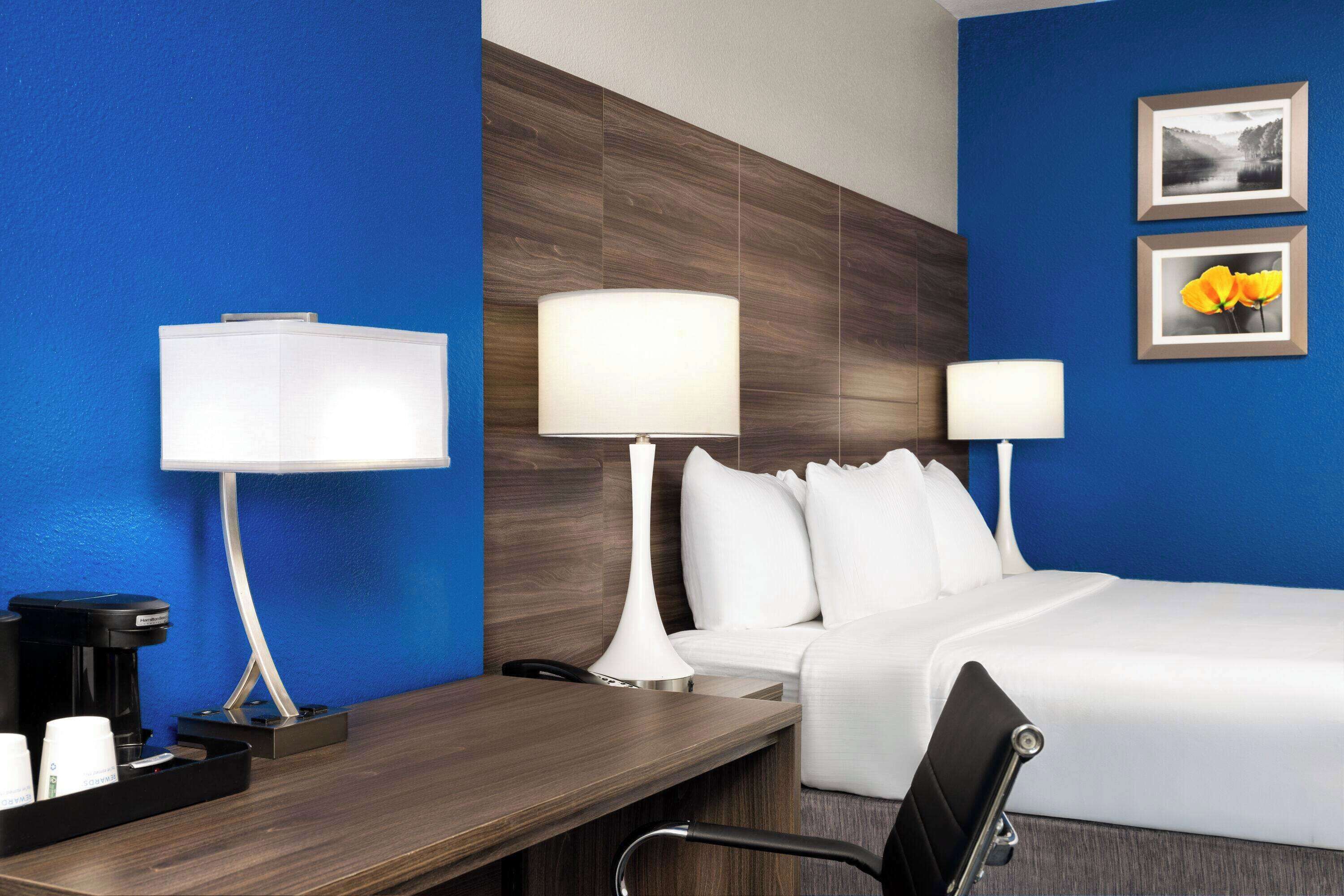 A hotel room with a bed, two bedside lamps, a desk with a coffee maker, and a chair.