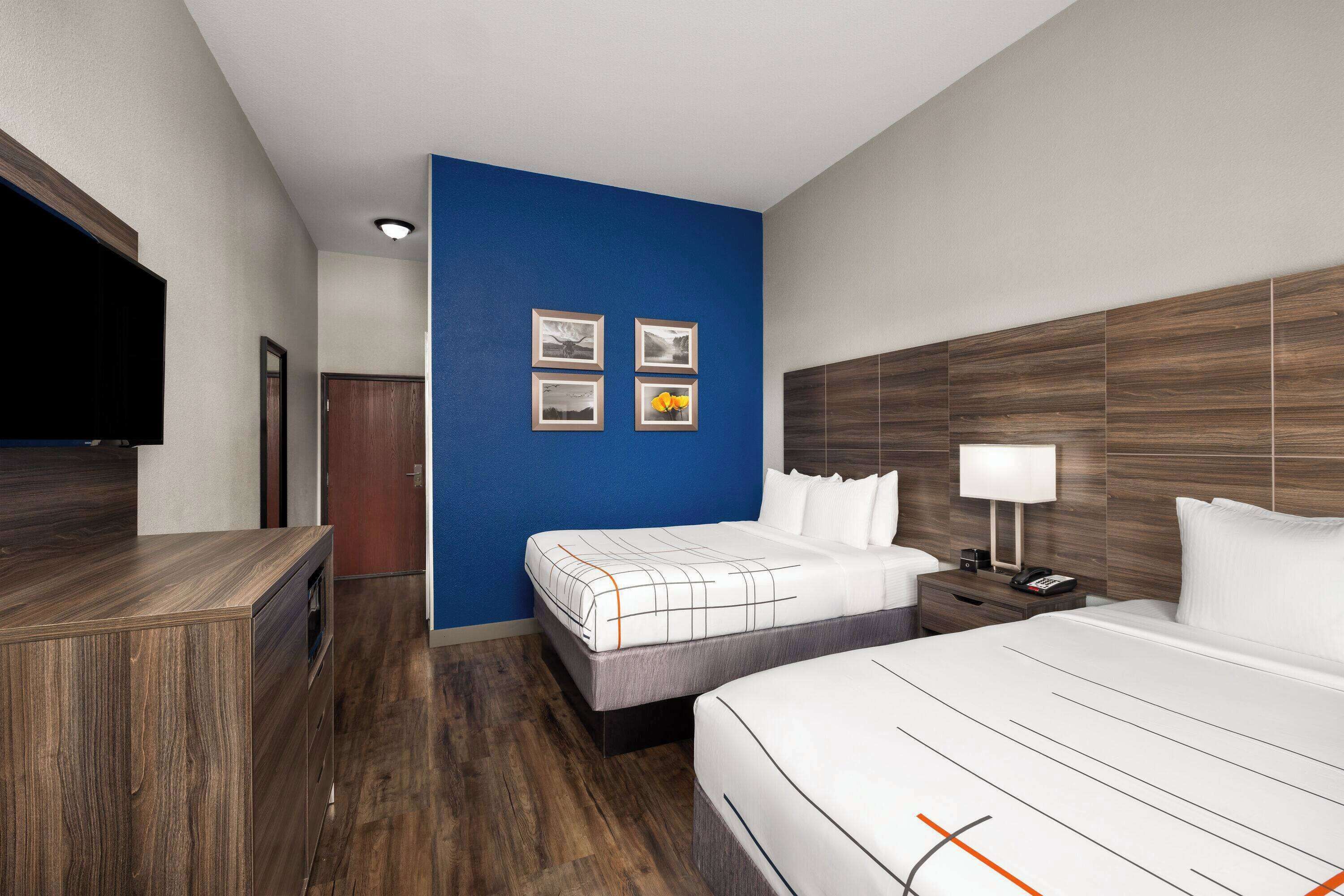 A hotel room with a wooden floor, a blue accent wall, a bed with white bedding, a nightstand with a lamp, and a flat-screen TV mounted on the wall.