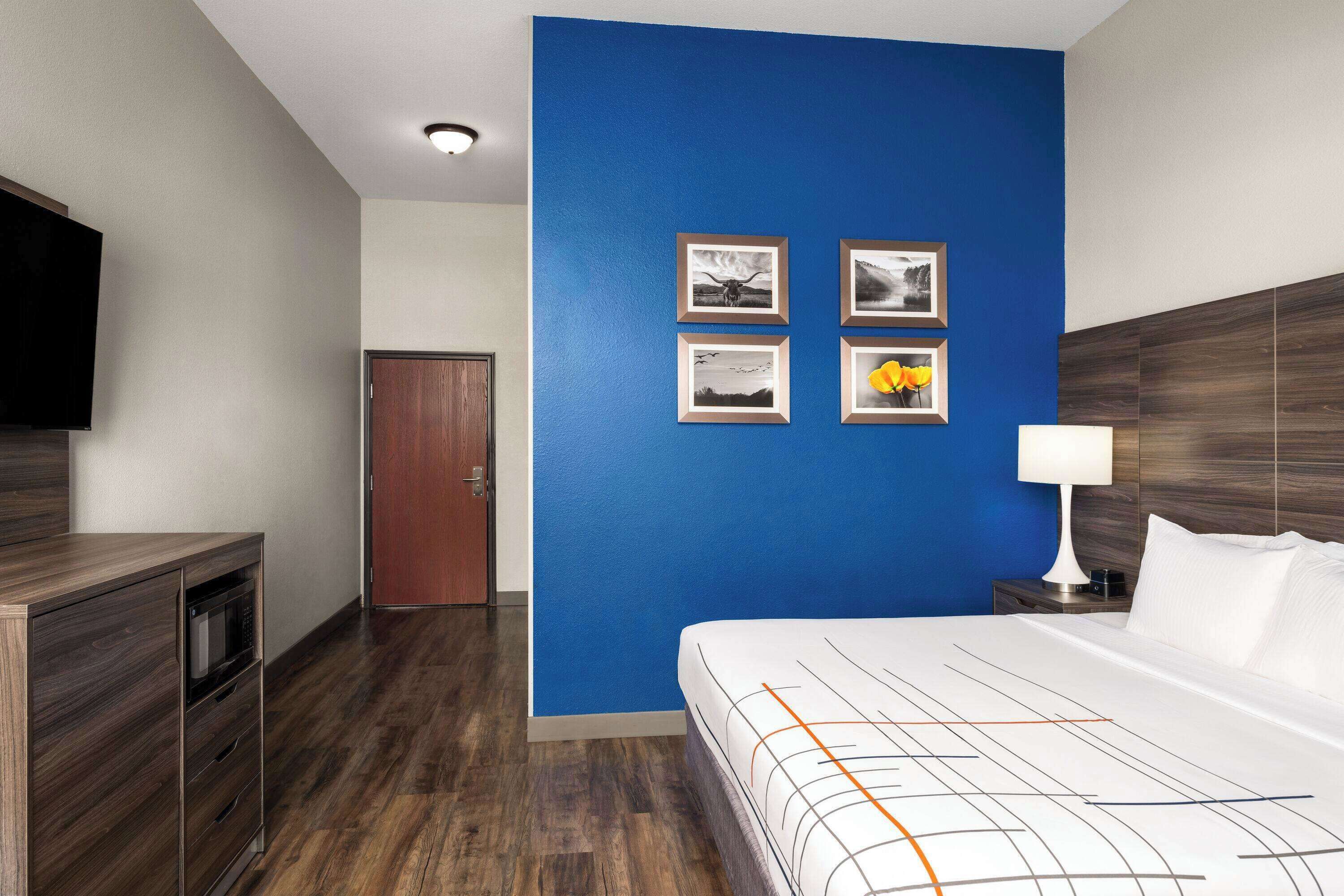 Habitació d'hotel amb una paret d'accent blau, un llit amb llençols blancs i taronges, un taulell de fusta amb microones i un televisor de pantalla plana penjat a la paret.