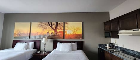 Een hotelkamer met twee bedden, een groot schilderij van een zonsondergang, een kitchenette met een magnetron en spoelbak, en houten kastjes.
