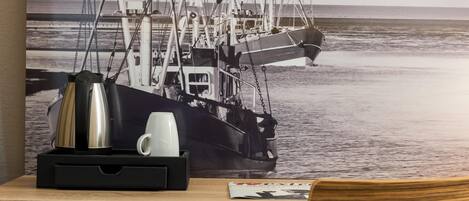 Un escritorio con una cafetera, taza y una revista, con un cartel de un barco en la pared.