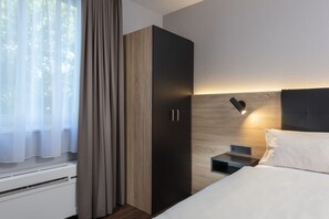 Une chambre à coucher moderne, dotée d’une grande armoire, d’une table de chevet avec une lampe et offrant une vue à travers des rideaux légers.