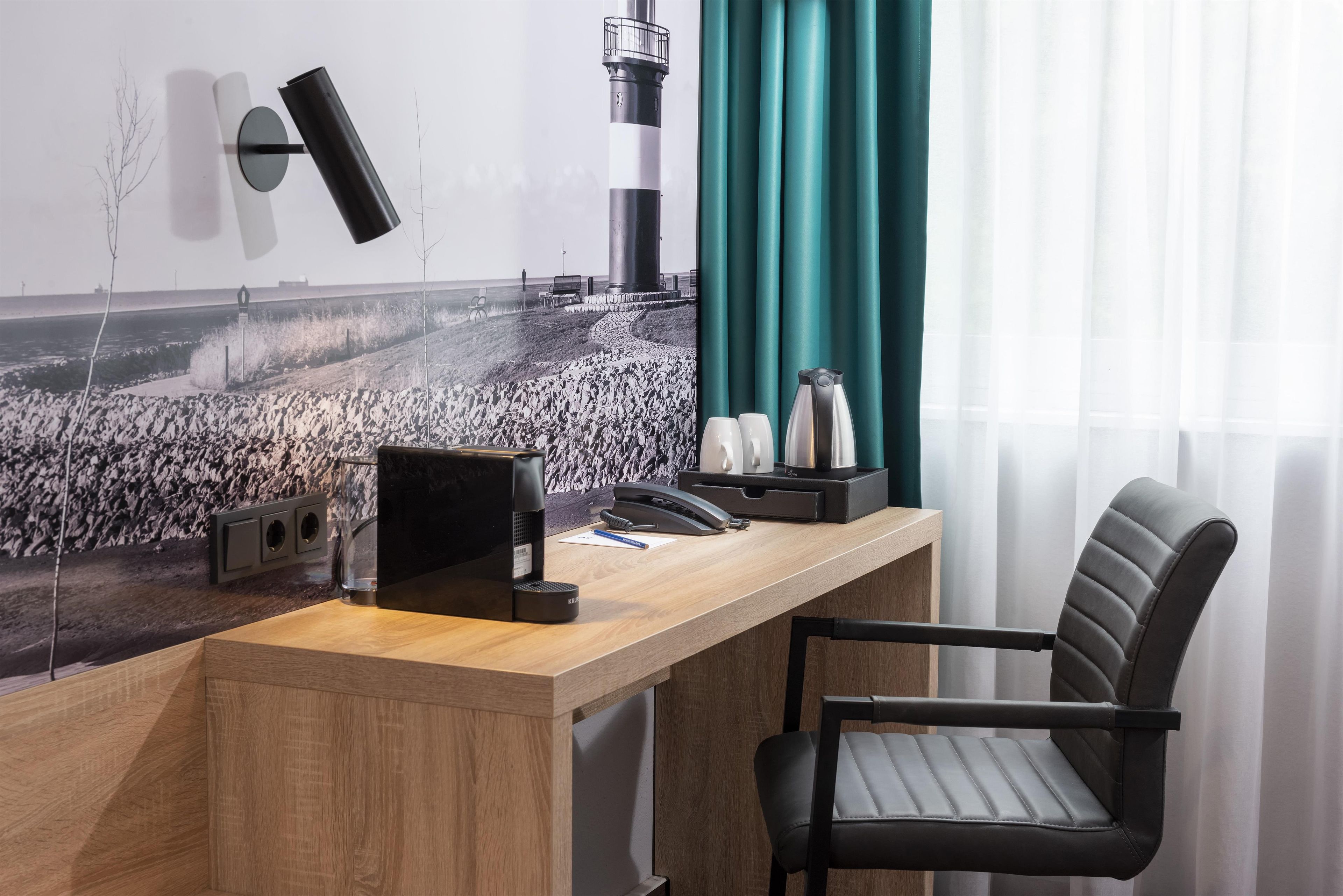 Ein Hotelzimmer mit einem Holztisch, einem schwarzen Bürostuhl, einer Wandlampe, einem Telefon und einer Kaffeemaschine.