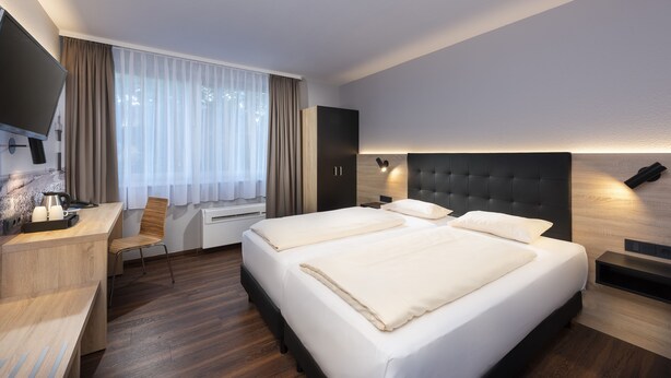 Una habitación de hotel moderna con una cama grande, un escritorio con silla, un televisor de pantalla plana y un ventanal con cortinas.