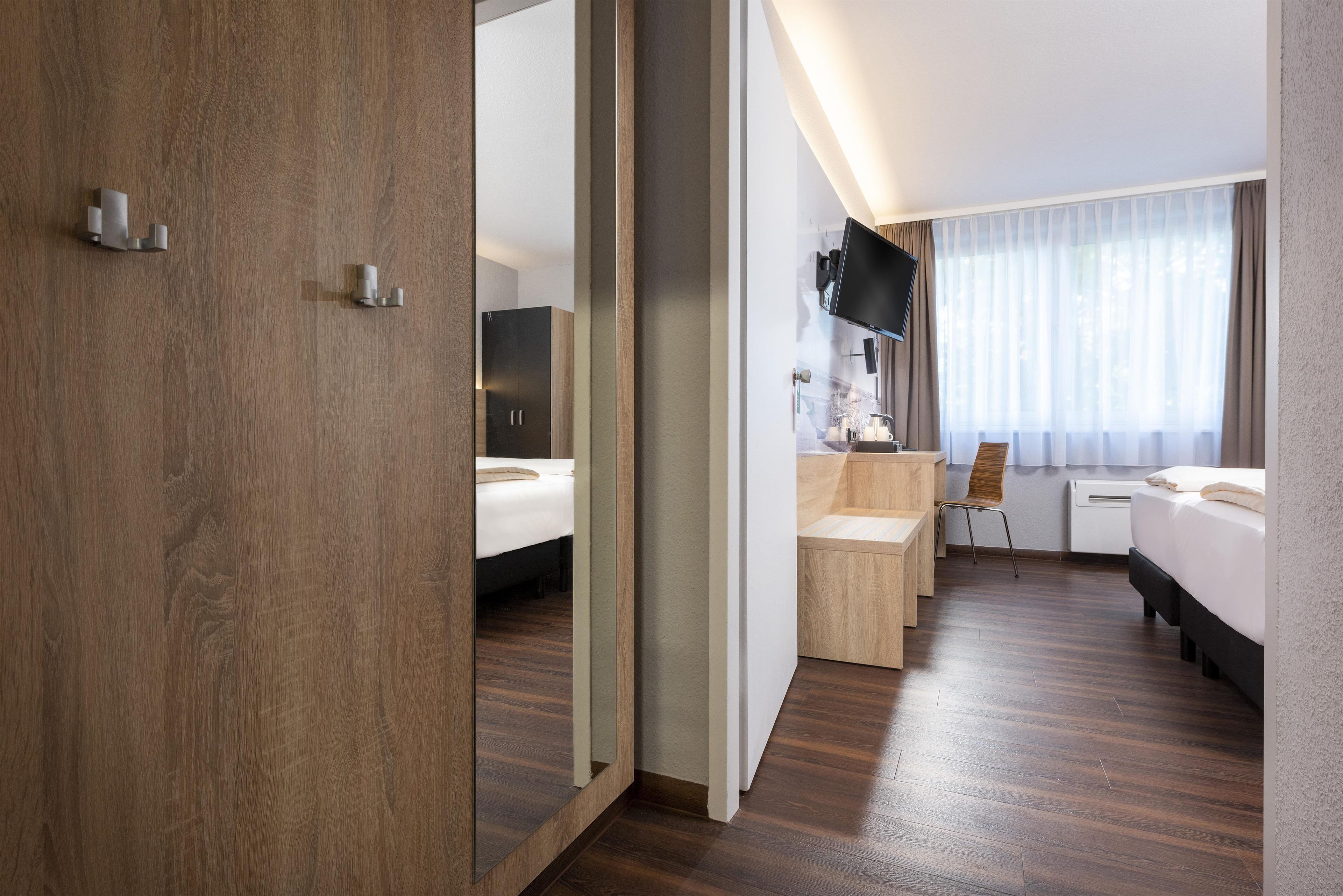 A hotel room with a wooden floor, a bed, a desk with a chair, a TV, and a window with curtains.