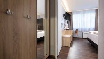 A hotel room with a wooden floor, a bed, a desk with a chair, a TV, and a window with curtains.