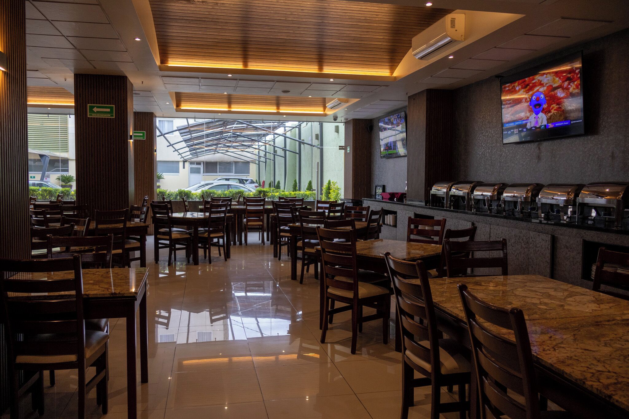 A restaurant with multiple tables and chairs, a counter with a beverage dispenser, and a television screen on the wall.