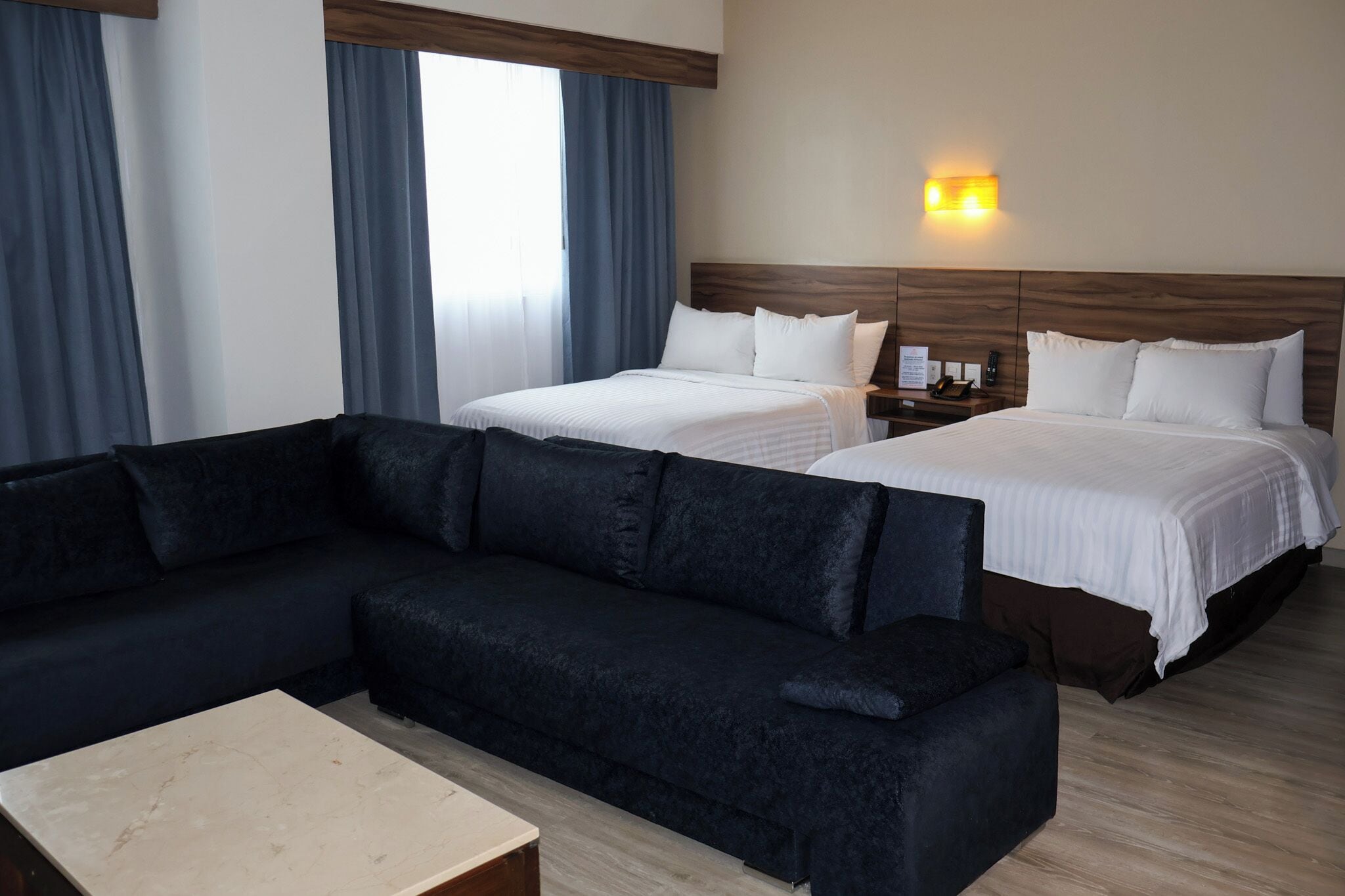 A hotel room with a dark-colored sofa, two beds with white bedding, a wooden headboard, a nightstand with a phone, and a wall-mounted light.