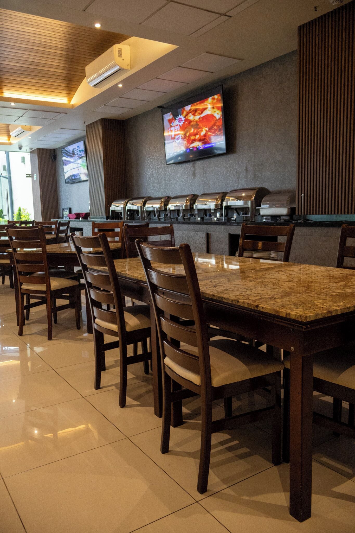 Un restaurant doté de nombreuses tables et chaises, d’un espace buffet avec des distributeurs de nourriture et d’un téléviseur fixé au mur.