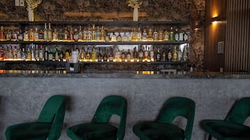 Baarissa on kiviseinä taustalla, useita eri alkoholijuomapulloja ja vihreät samettiset tuolit.