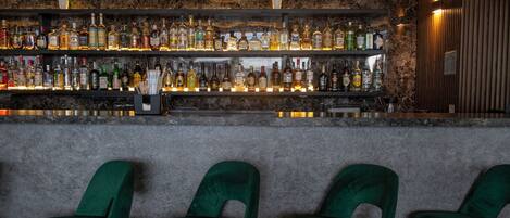 A bar with a stone wall background, multiple liquor bottles, and green velvet chairs.