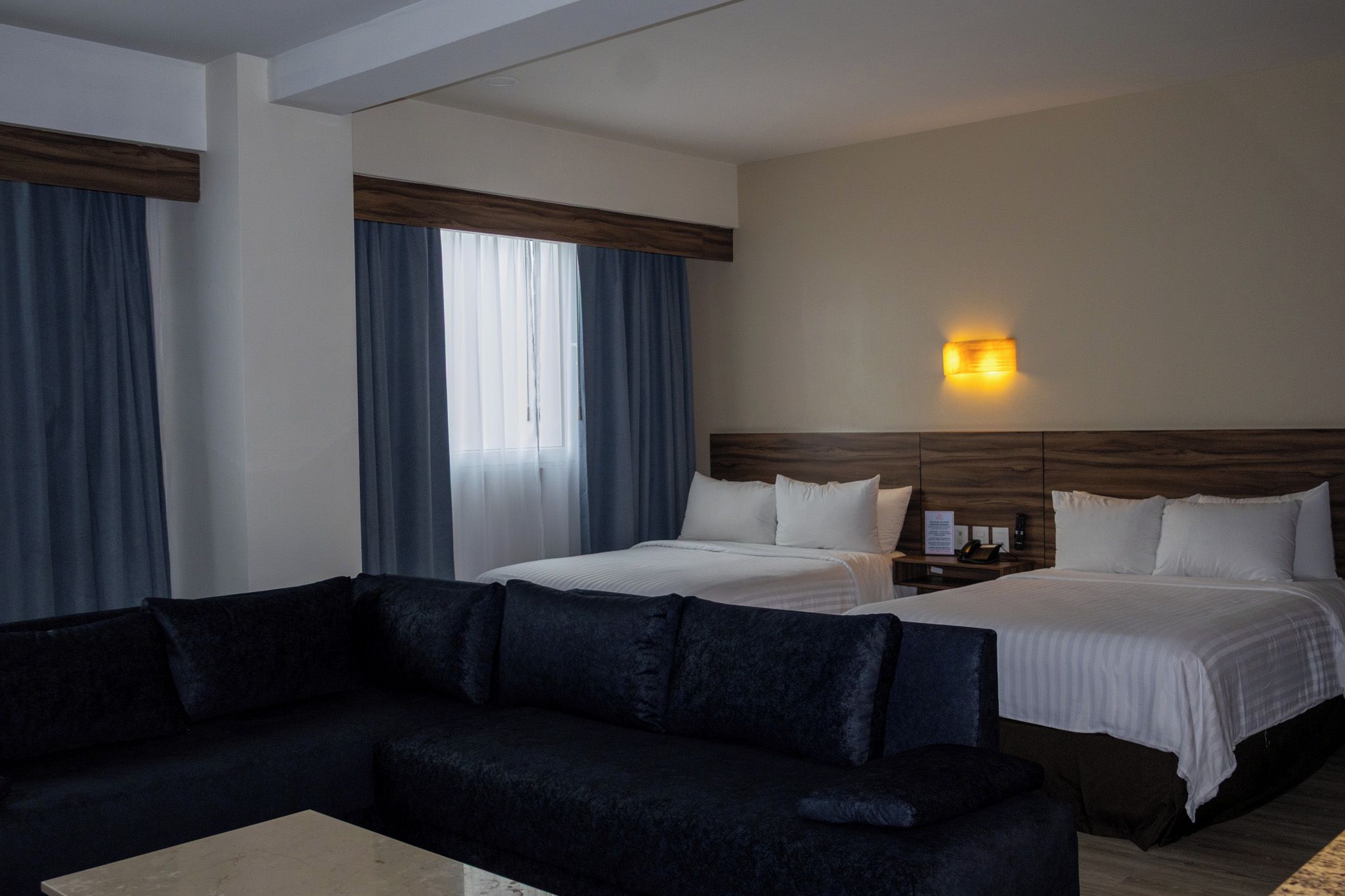 Habitación de hotel con un sofá de color oscuro, dos camas con sábanas blancas, cabeceros de madera, una mesita de noche con lámpara y una ventana con cortinas azules.
