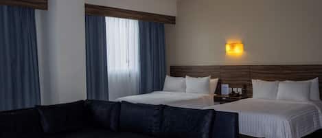 Habitación de hotel con un sofá de color oscuro, dos camas con sábanas blancas, cabeceros de madera, una mesita de noche con lámpara y una ventana con cortinas azules.