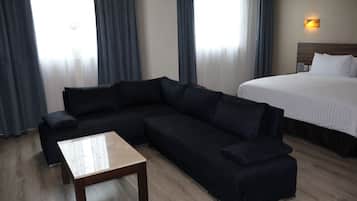 Habitación de hotel con sofá oscuro, una mesita de madera, cama con ropa blanca y una lámpara fijada a la pared.