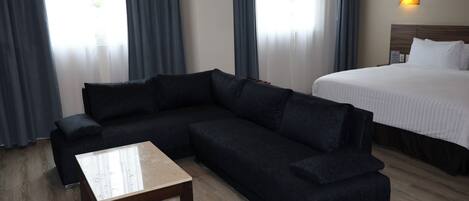 Habitación de hotel con sofá oscuro, una mesita de madera, cama con ropa blanca y una lámpara fijada a la pared.