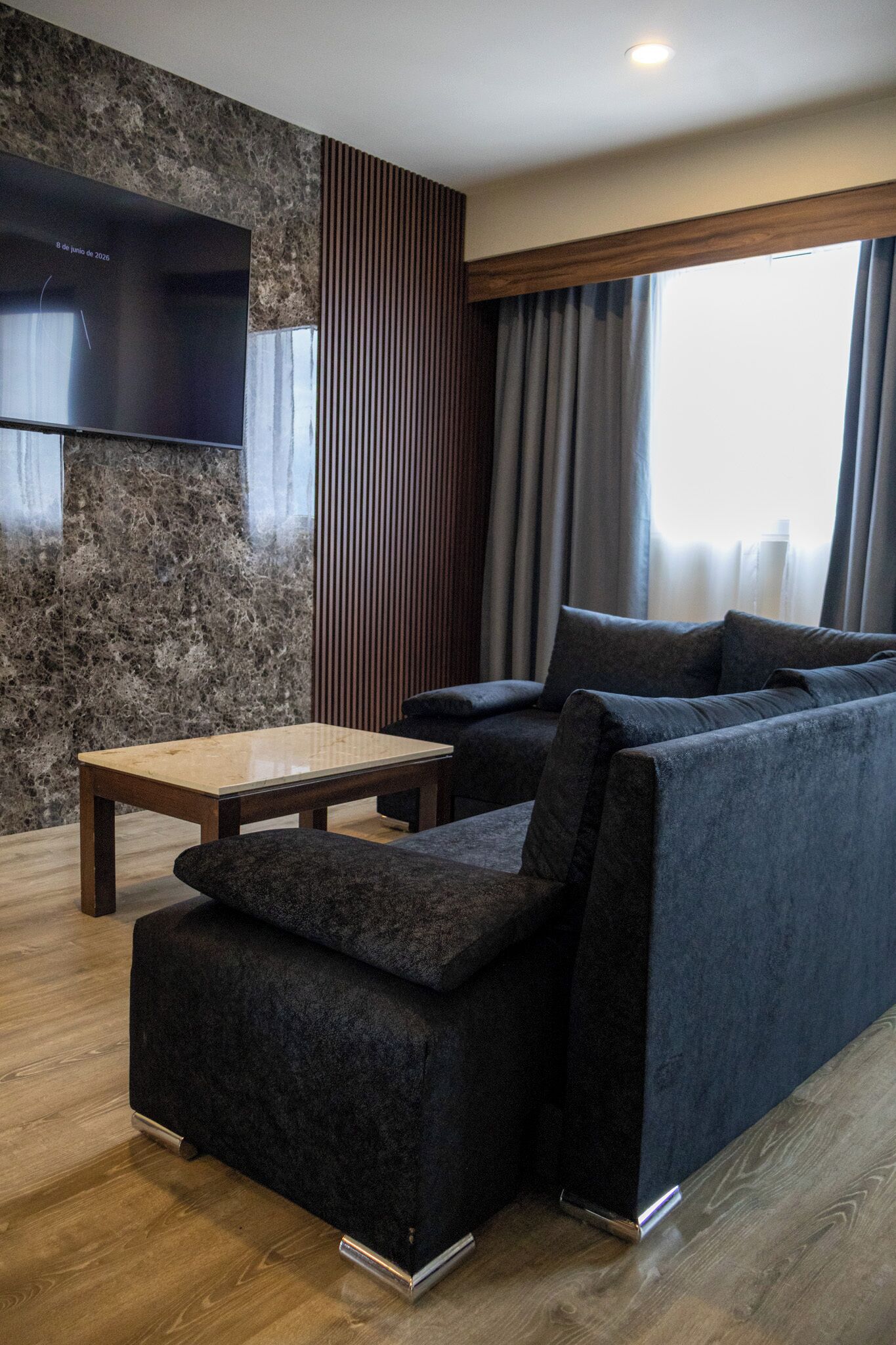 A modern living room with a sectional sofa, a flat-screen TV mounted on a textured wall, a wooden coffee table, and a large window with curtains.