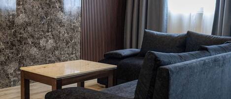 Ein modernes Wohnzimmer mit einem Ecksofa, einem flachbildfernseher, der an einer strukturierten Wand befestigt ist, einem Holztisch und einem großen Fenster mit Vorhängen.