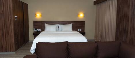 Habitación de hotel con una cama grande, un armario de madera, un sofá marrón y dos lámparas fijadas a la pared.