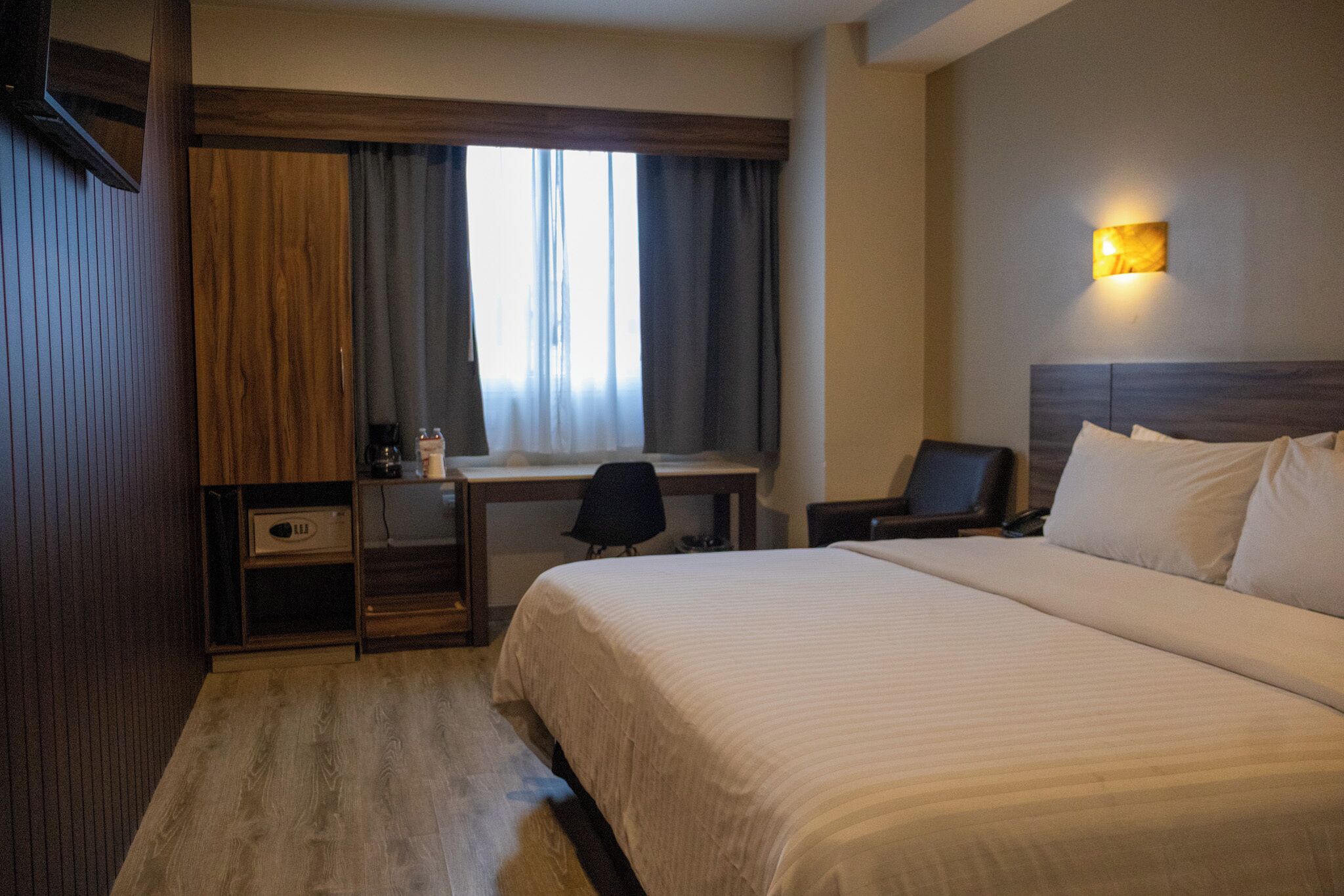 A hotel room with a bed, desk, chair, and a wall-mounted light fixture.