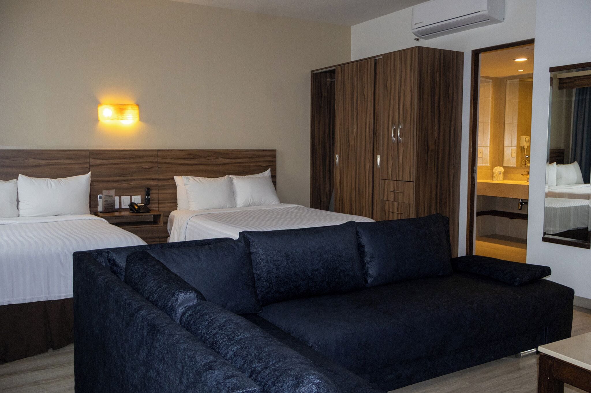 A hotel room with two beds, a dark sofa, a wardrobe, and a bathroom visible through an open door.
