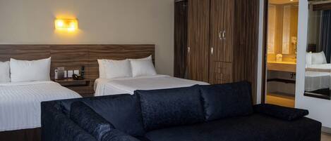A hotel room with two beds, a dark sofa, a wardrobe, and a bathroom visible through an open door.