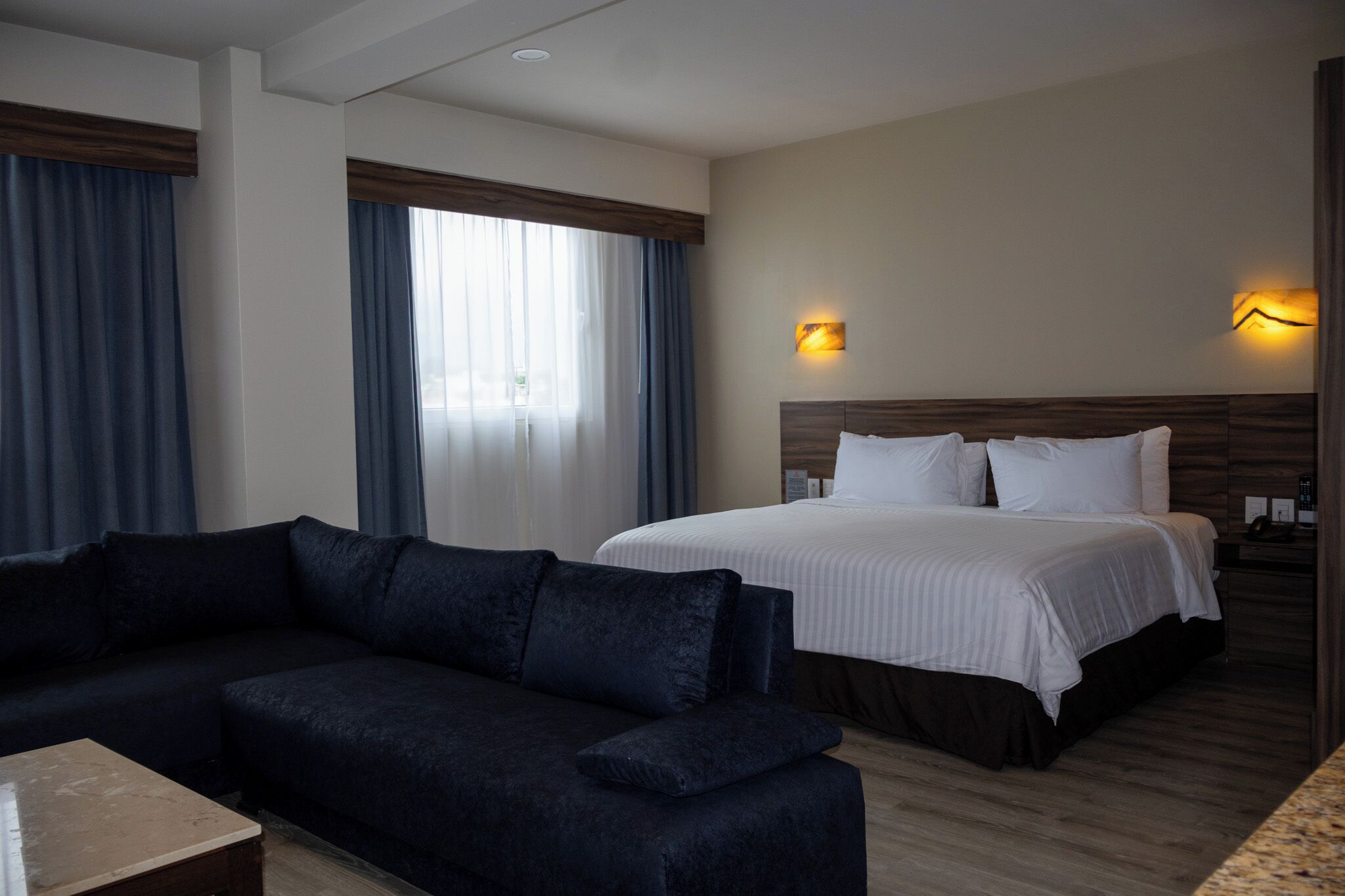 A hotel room with a bed, a dark-colored sofa, a small table, and two wall-mounted light fixtures.