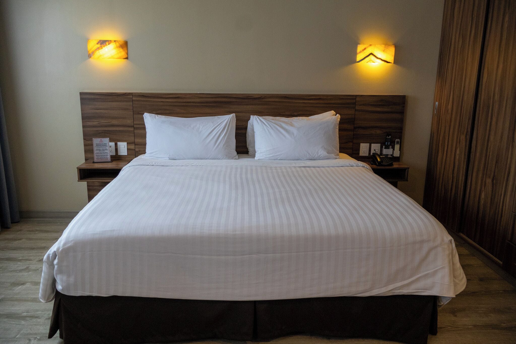 Una cama bien hecha con cabecera, dos mesitas de noche con lámparas y suelo de madera.