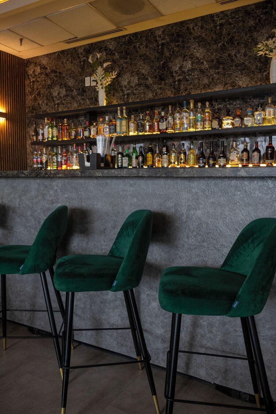 Een bar met een marmeren wand, groene barkrukken en diverse flessen drank.