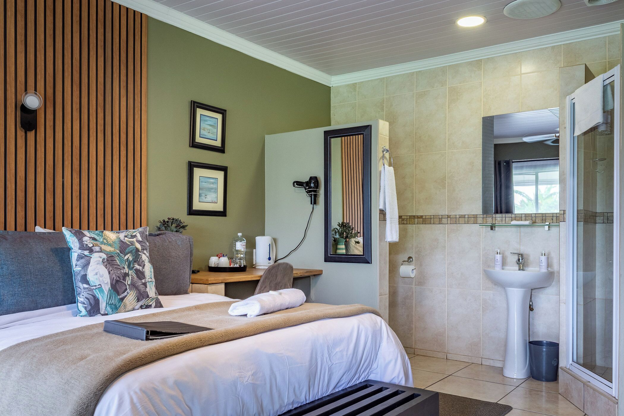 A hotel room with a bed, a bedside table with a hairdryer, a mirror, a sink, and a shower.