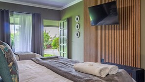 A modern hotel room with a large bed, a flat-screen TV mounted on a wooden panel wall, and a view of the outdoors through a glass door. - Sunrock Guesthouse (Kempton Park)