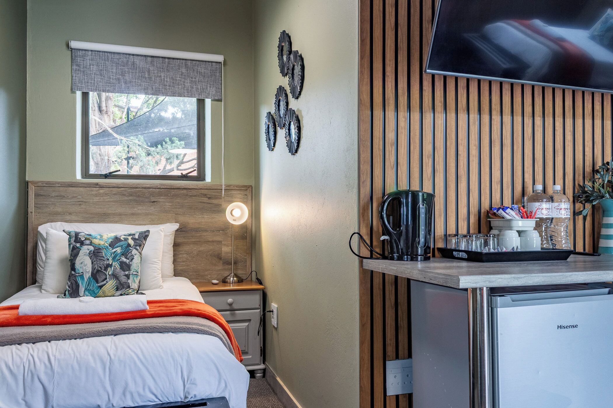 A hotel room with a bed, bedside lamp, a small table with a coffee maker, and a TV mounted on the wall.