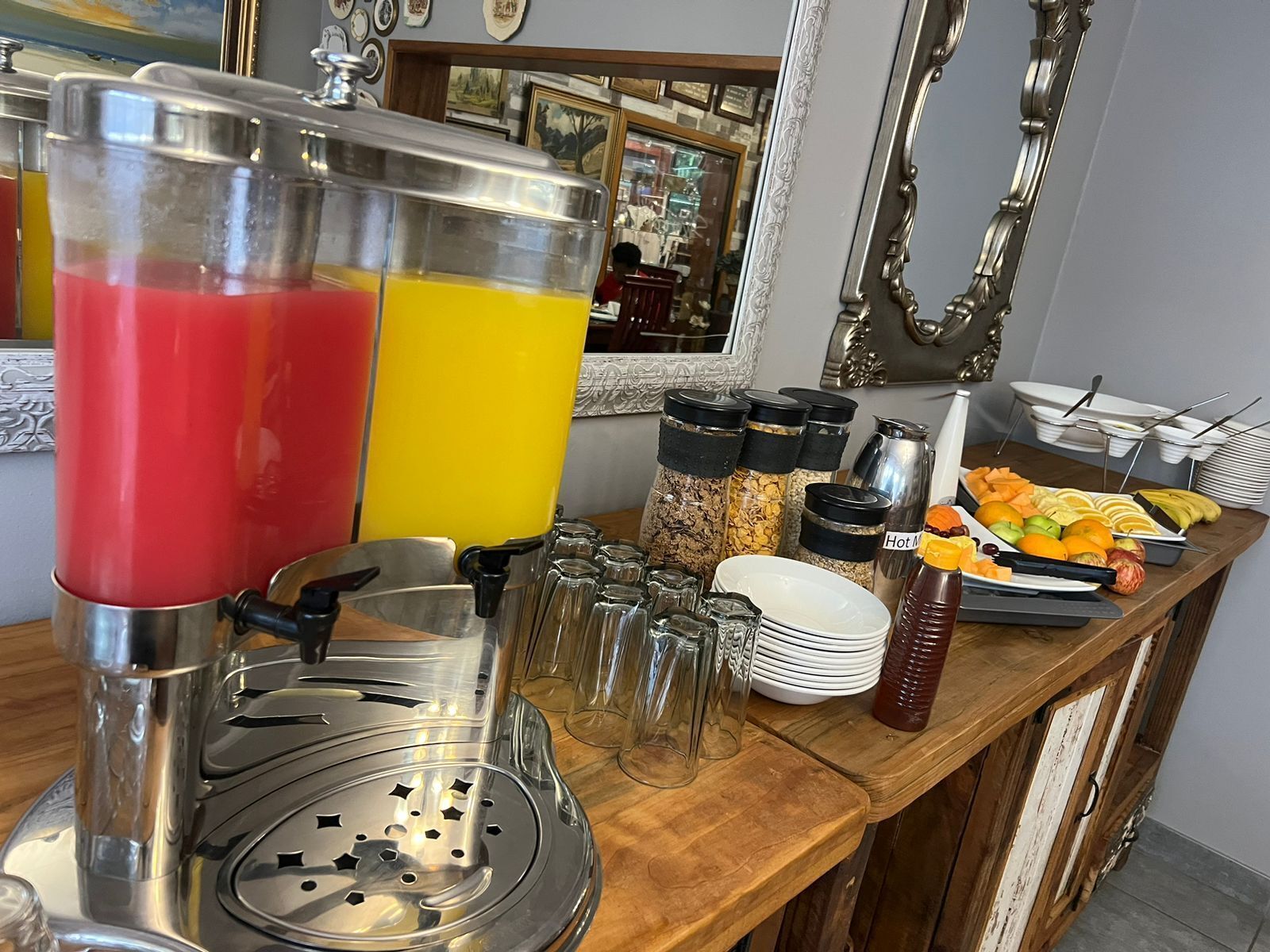 Una barra de desayuno con dispensador de zumo de frutas, frascos de condimentos y una bandeja de fruta.