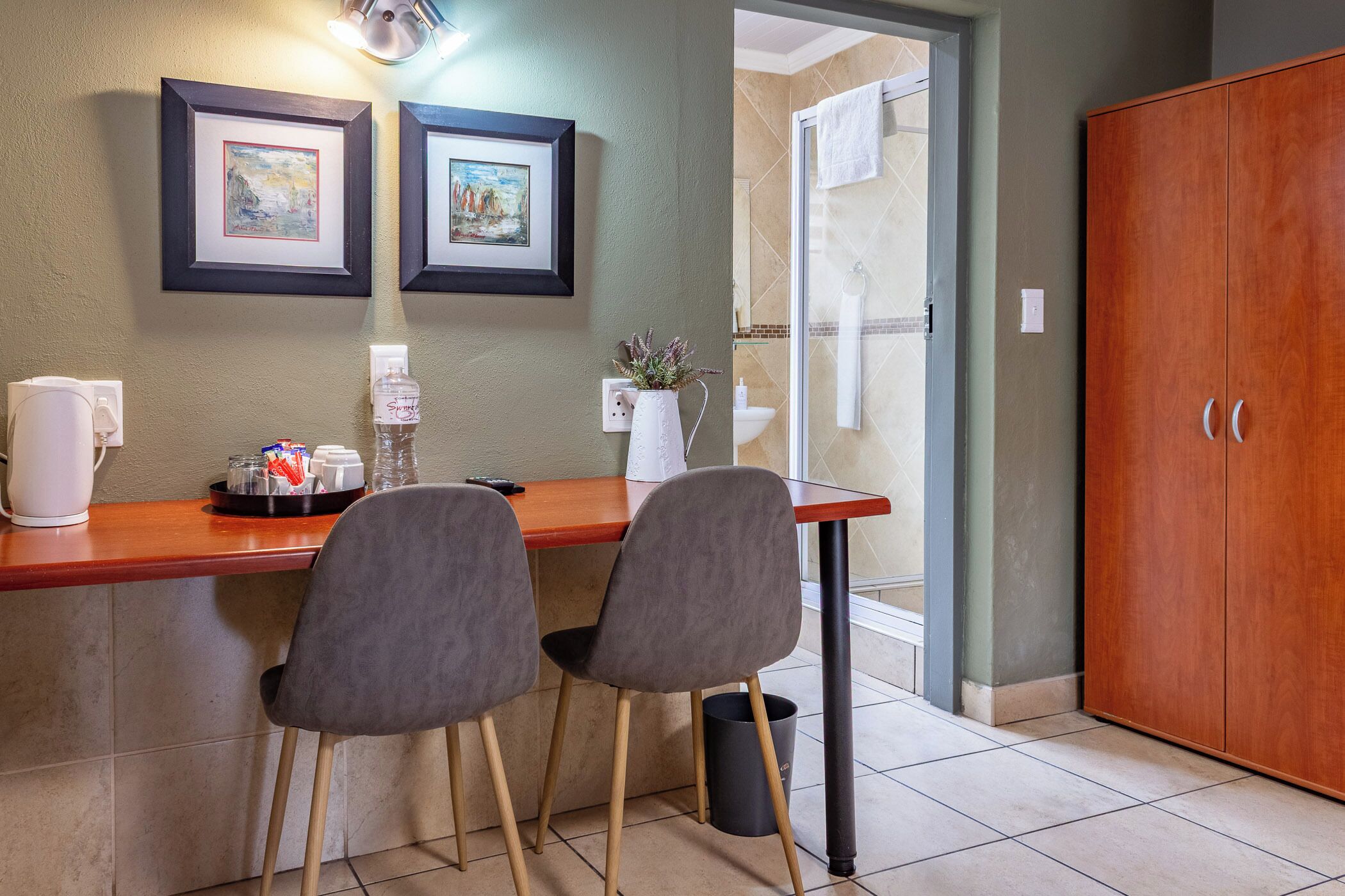 A small kitchenette with two chairs, a wooden table, framed pictures on the wall, and a view of the bathroom.