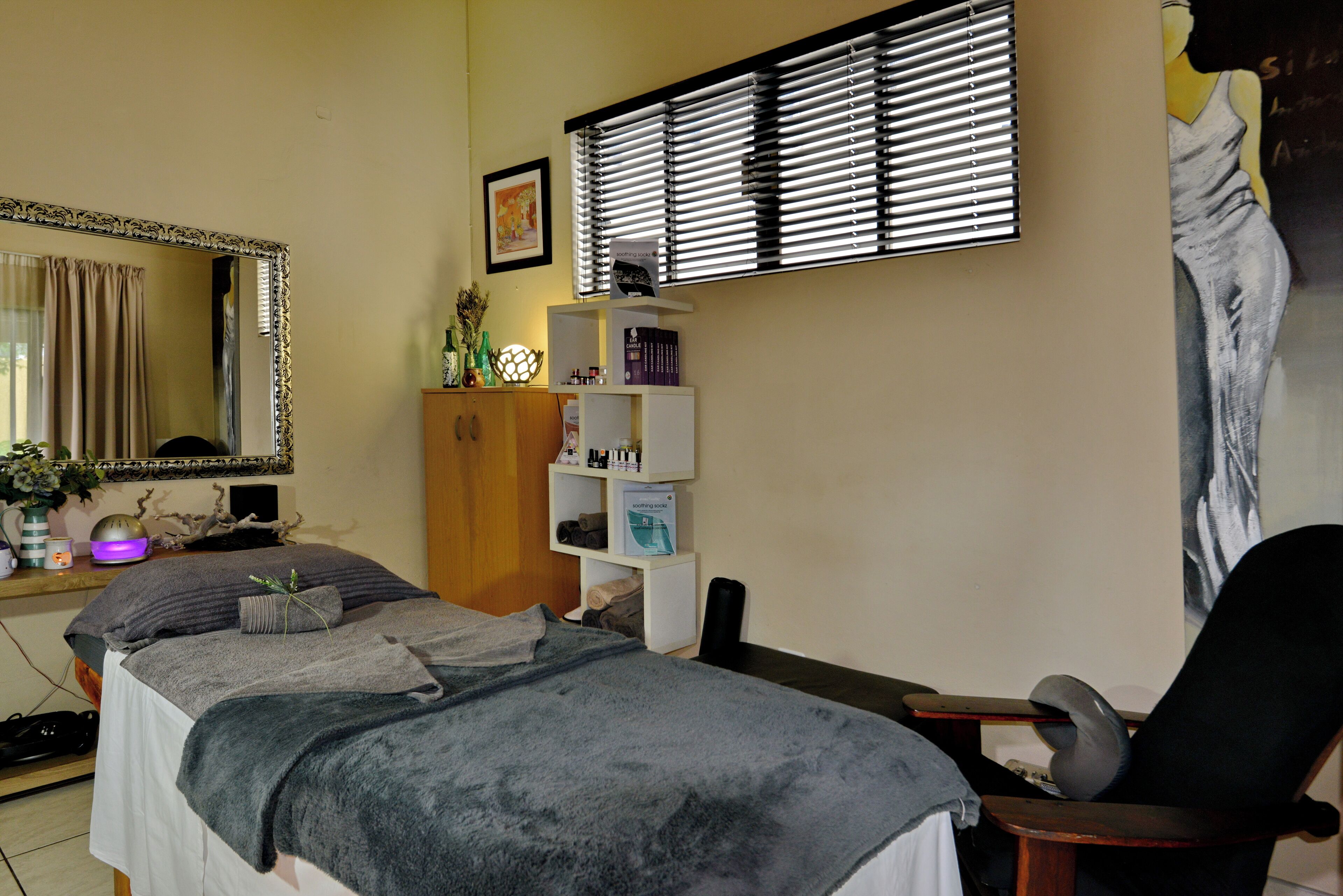 A bedroom with a bed, a chair, a shelf with books, a mirror, and a window with blinds.