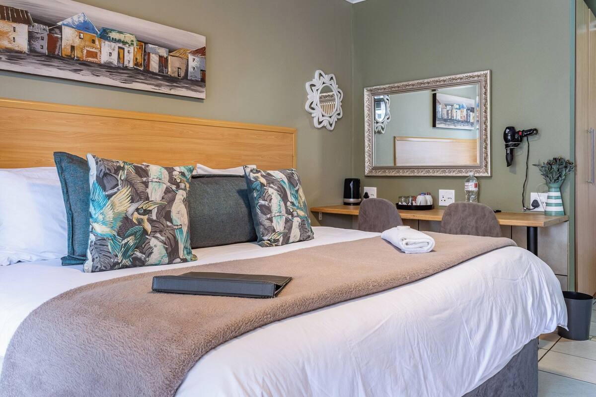 A neatly made bed with a brown blanket and decorative pillows, a wooden headboard, a desk with a chair, a mirror, and a wall-mounted hairdryer.