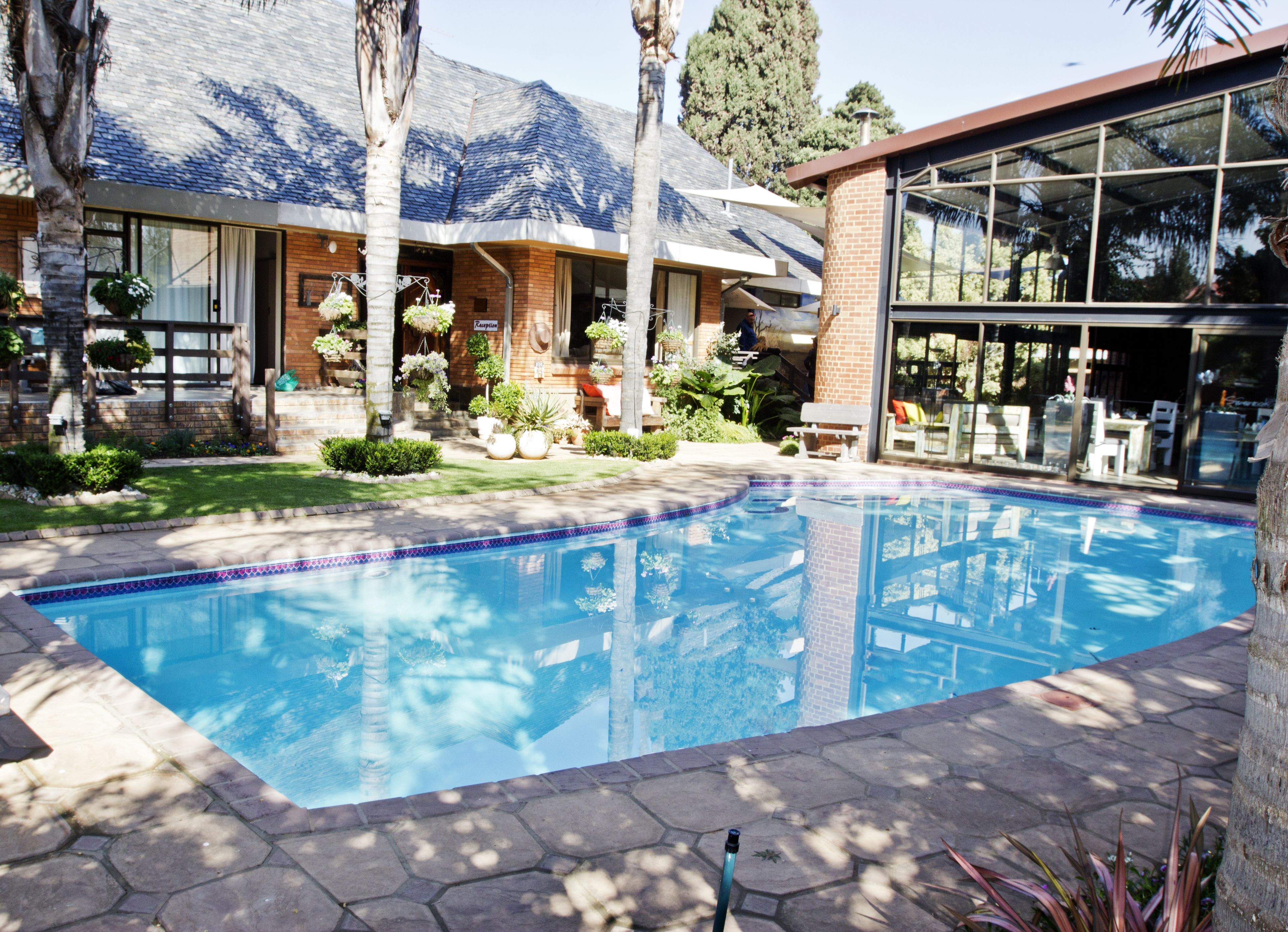 Una piscina rodeada por un jardín ajardinado, un edificio de ladrillo con grandes ventanales y un patio con mobiliario exterior.