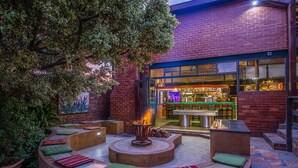 An outdoor seating area with a fire pit, cushioned benches, and a bar visible through the glass doors. - Sunrock Guesthouse (Kempton Park)