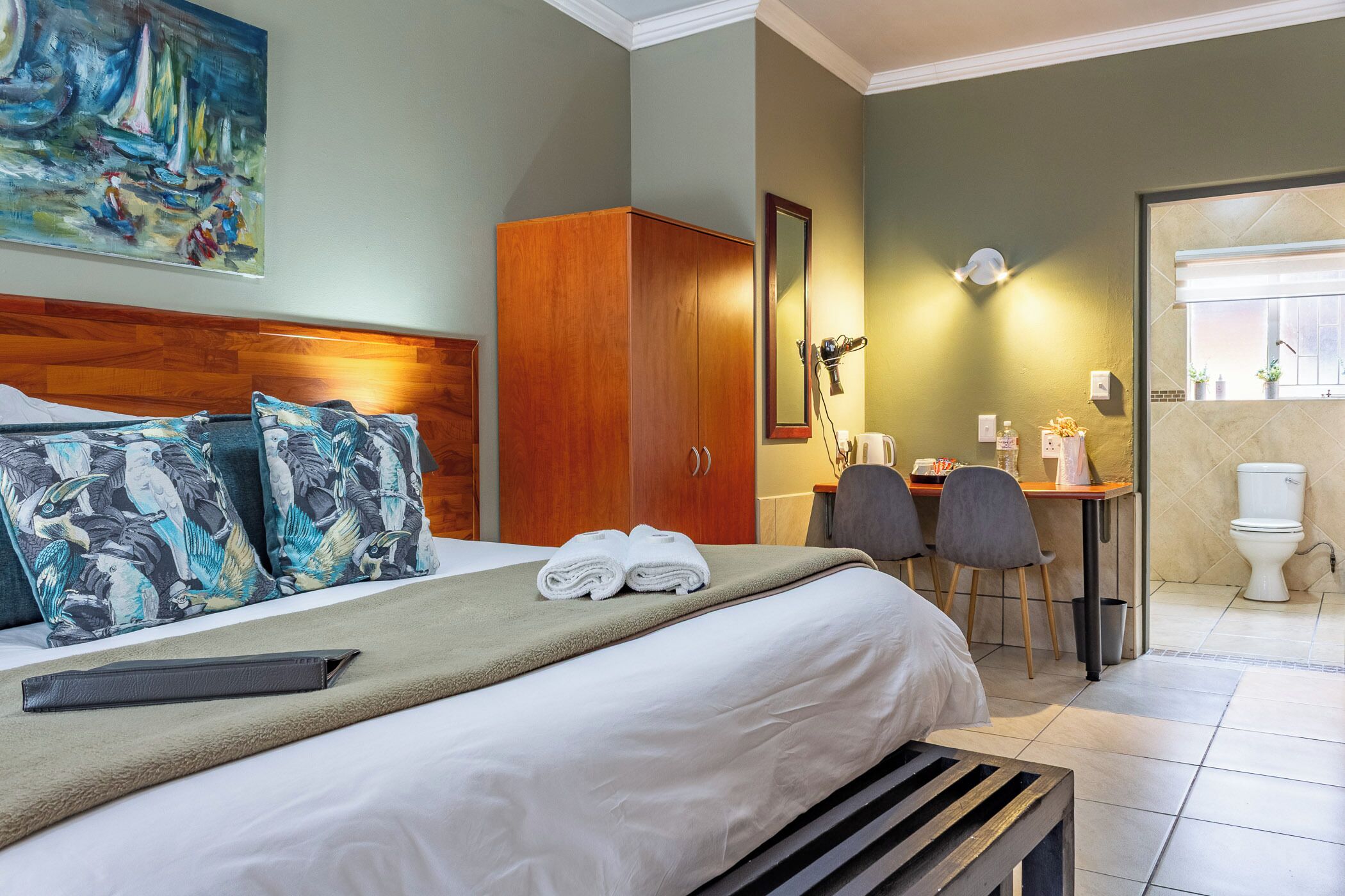 A hotel room with a bed, a wooden wardrobe, a desk with chairs, and a bathroom visible through an open door.