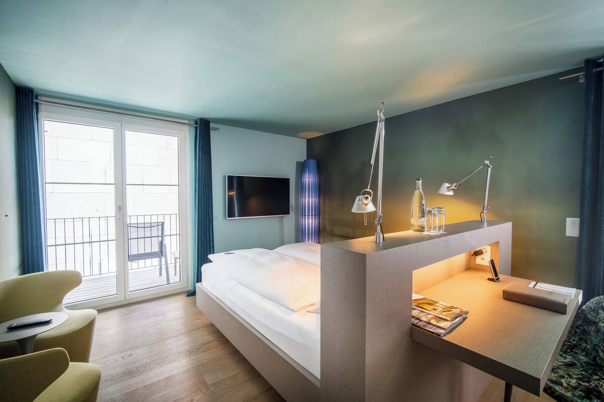 Ein modernes Hotelzimmer mit einem Bett, einem Schreibtisch mit Lampe, einem Sessel und einem Fernseher an der Wand. Es besitzt einen Balkon mit Sitzgelegenheit.
