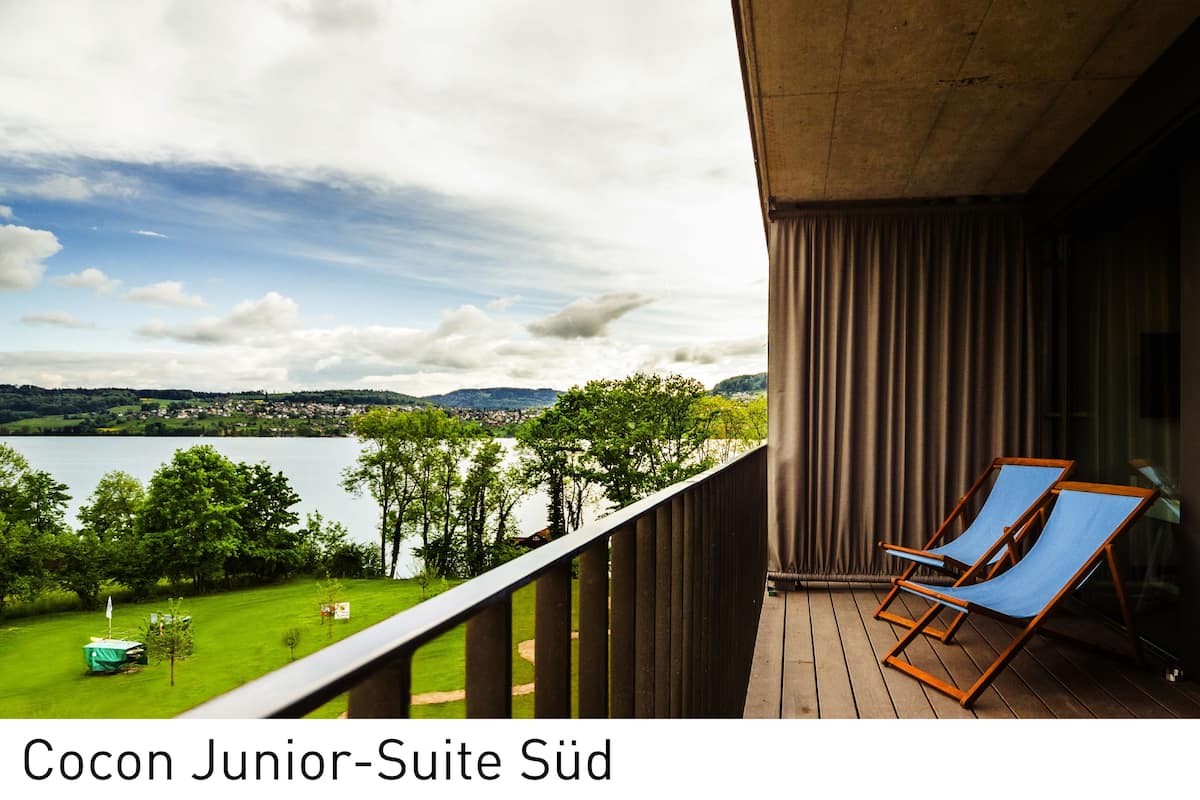 Ein Balkon mit zwei blauen Lounge-Stühlen, Blick auf einen See und eine entfernte Stadt.