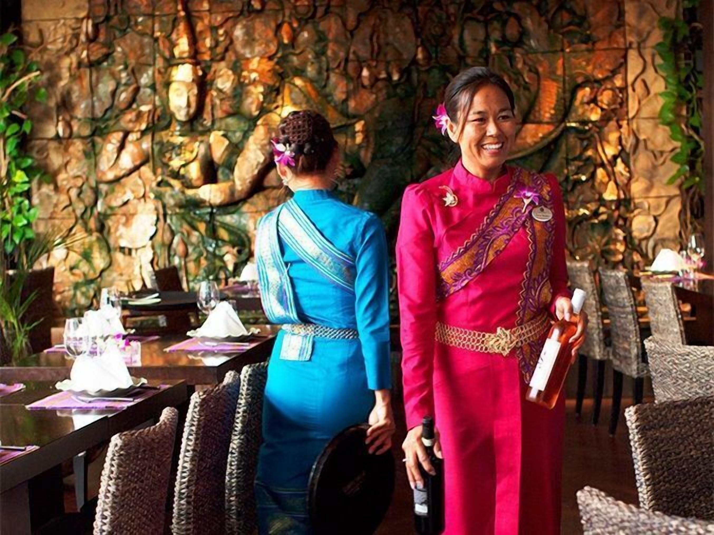 Two people in traditional attire standing in a restaurant with a stone wall background and wicker furniture.
