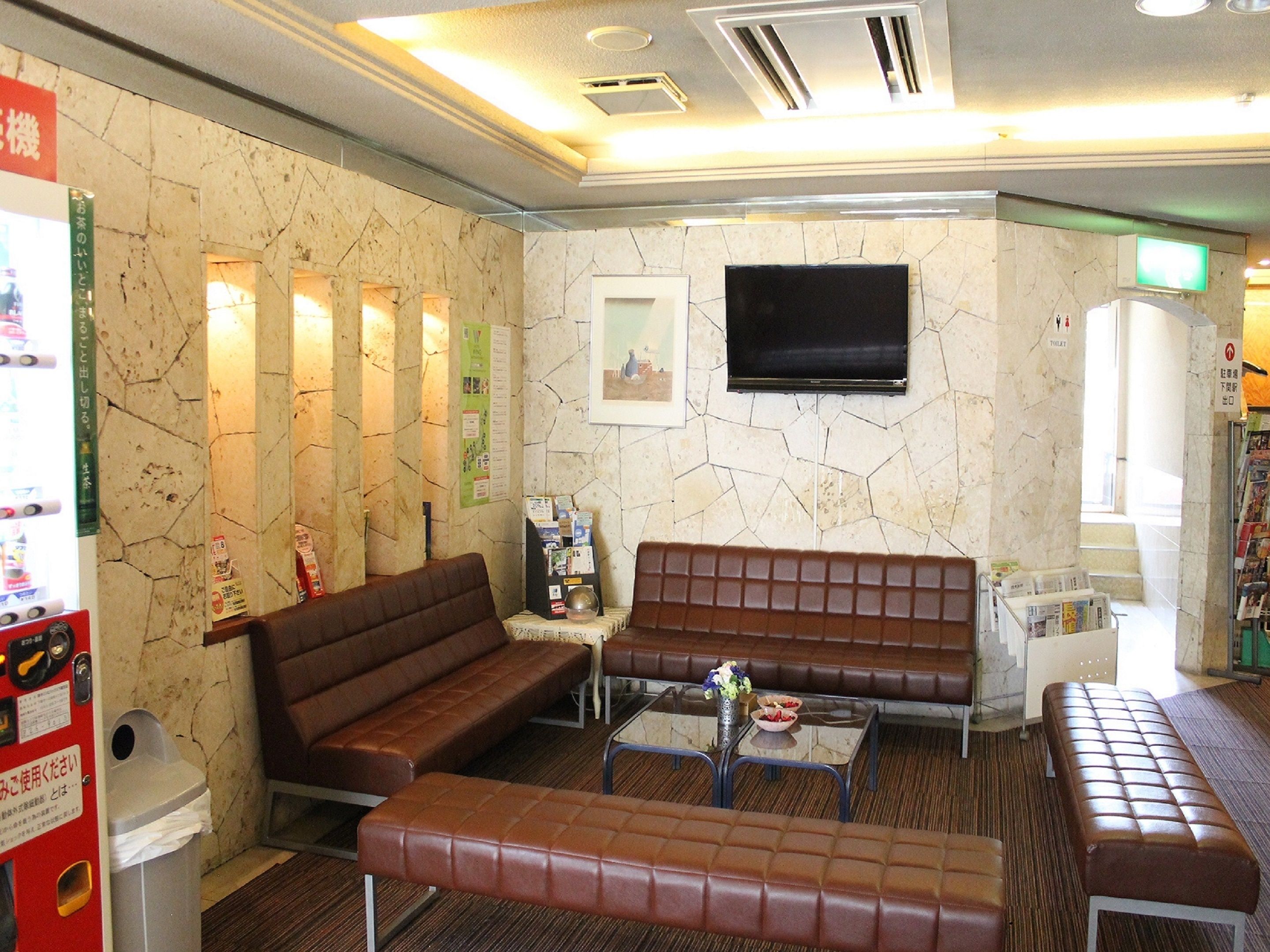 A lobby with a brown leather sofa, a small table with a vase, a trash bin, and a television mounted on the wall.