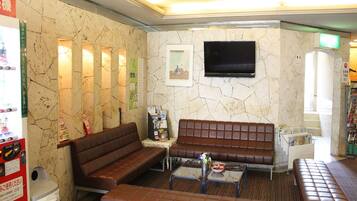 A lobby with a brown leather sofa, a small table with a vase, a trash bin, and a television mounted on the wall.