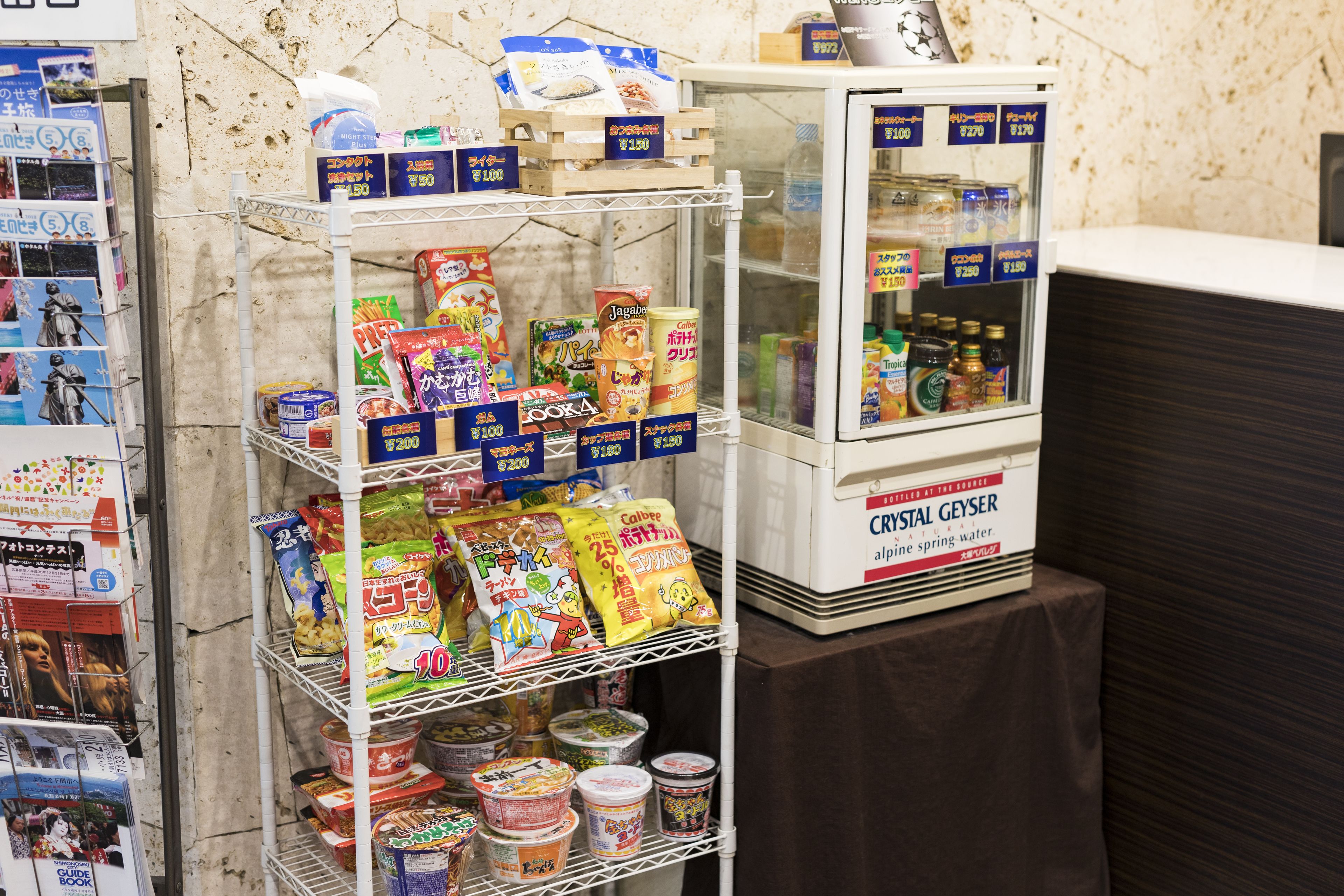 Un estante con refrigerios y bebidas, un refrigerador con refrescos, y una selección de revistas.