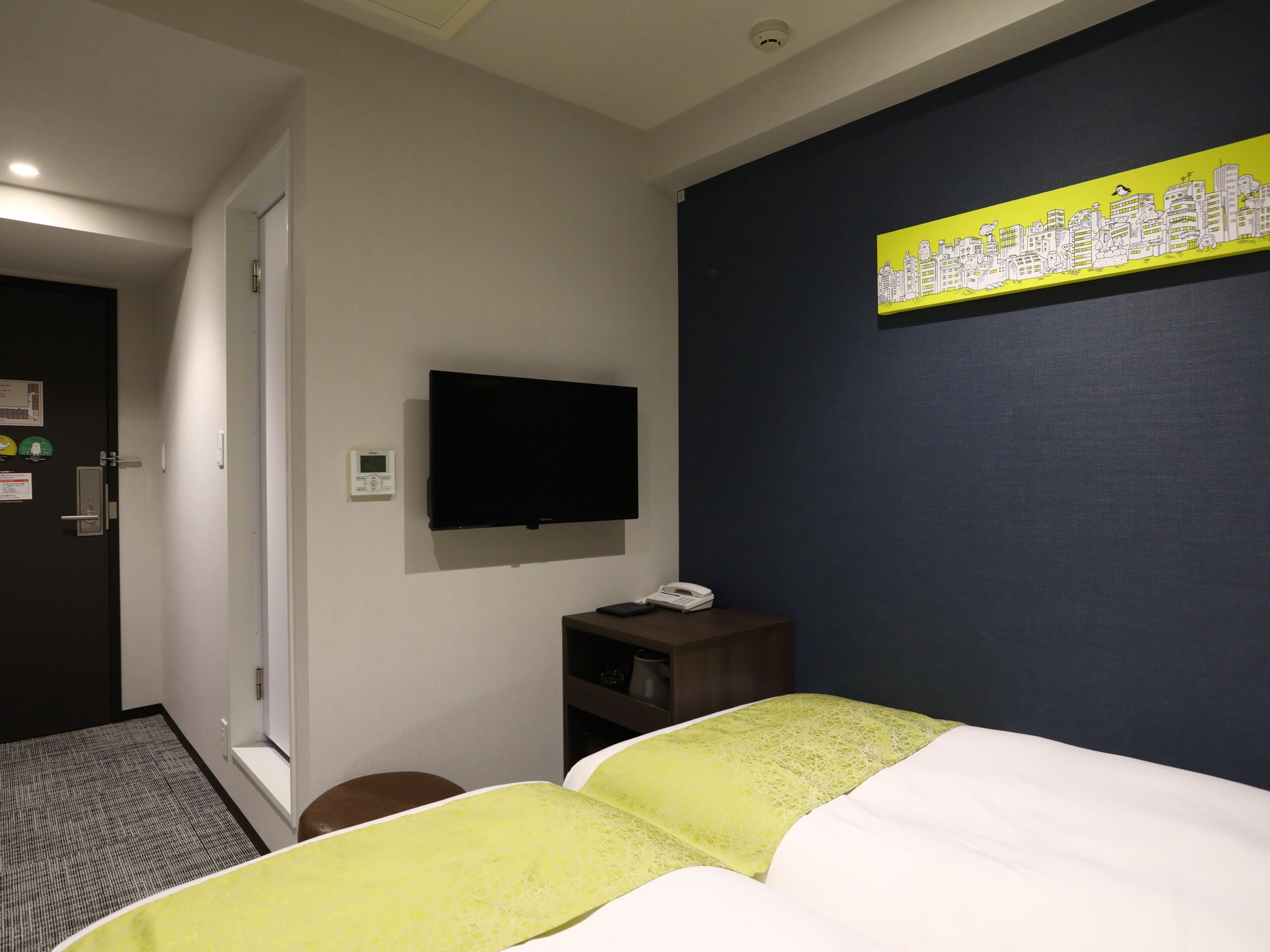 A hotel room with a bed, a flat-screen TV mounted on the wall, a bedside table with a phone, and a door with a handle.
