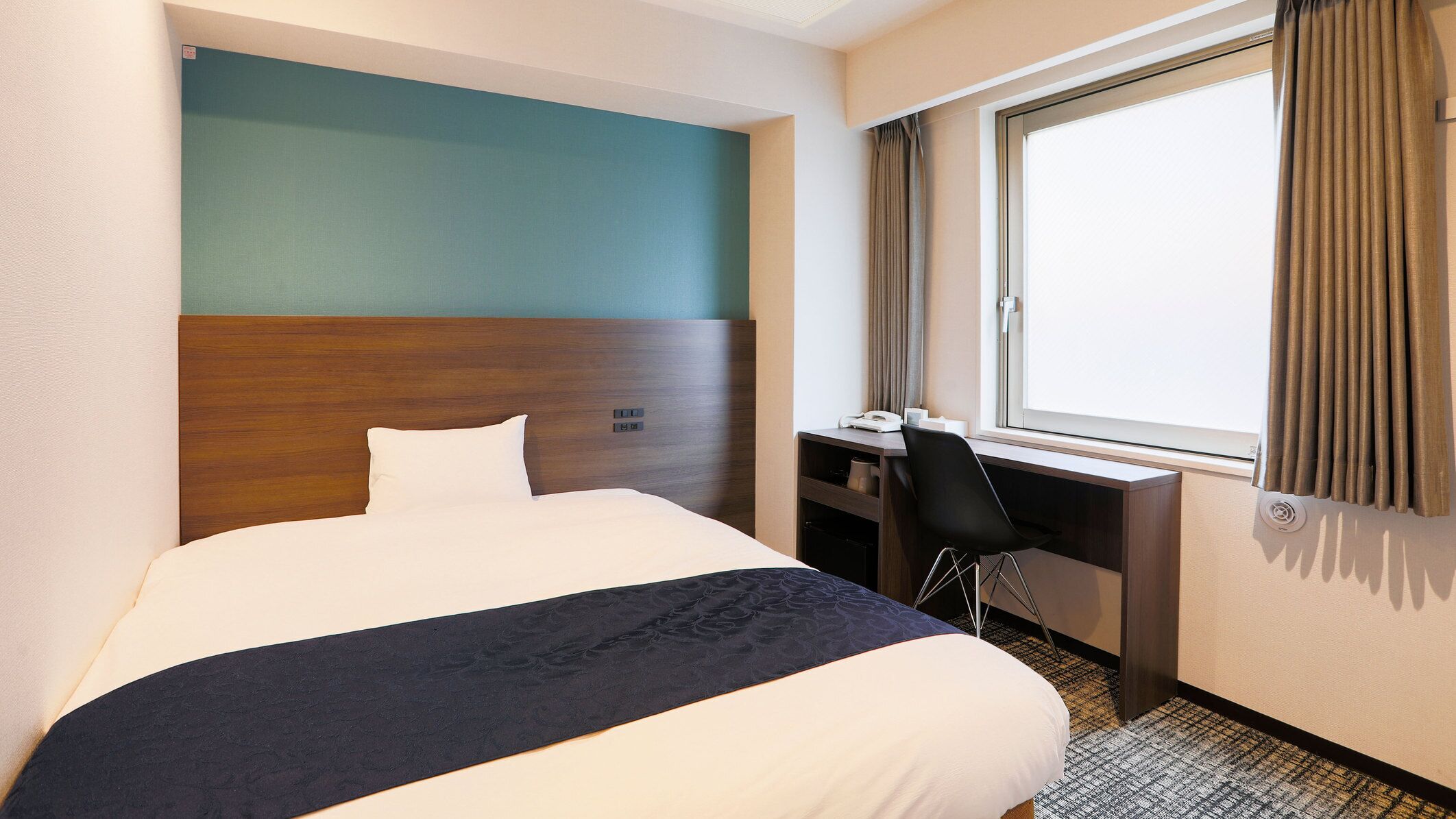 A hotel room with a bed, a desk, and a window with curtains.