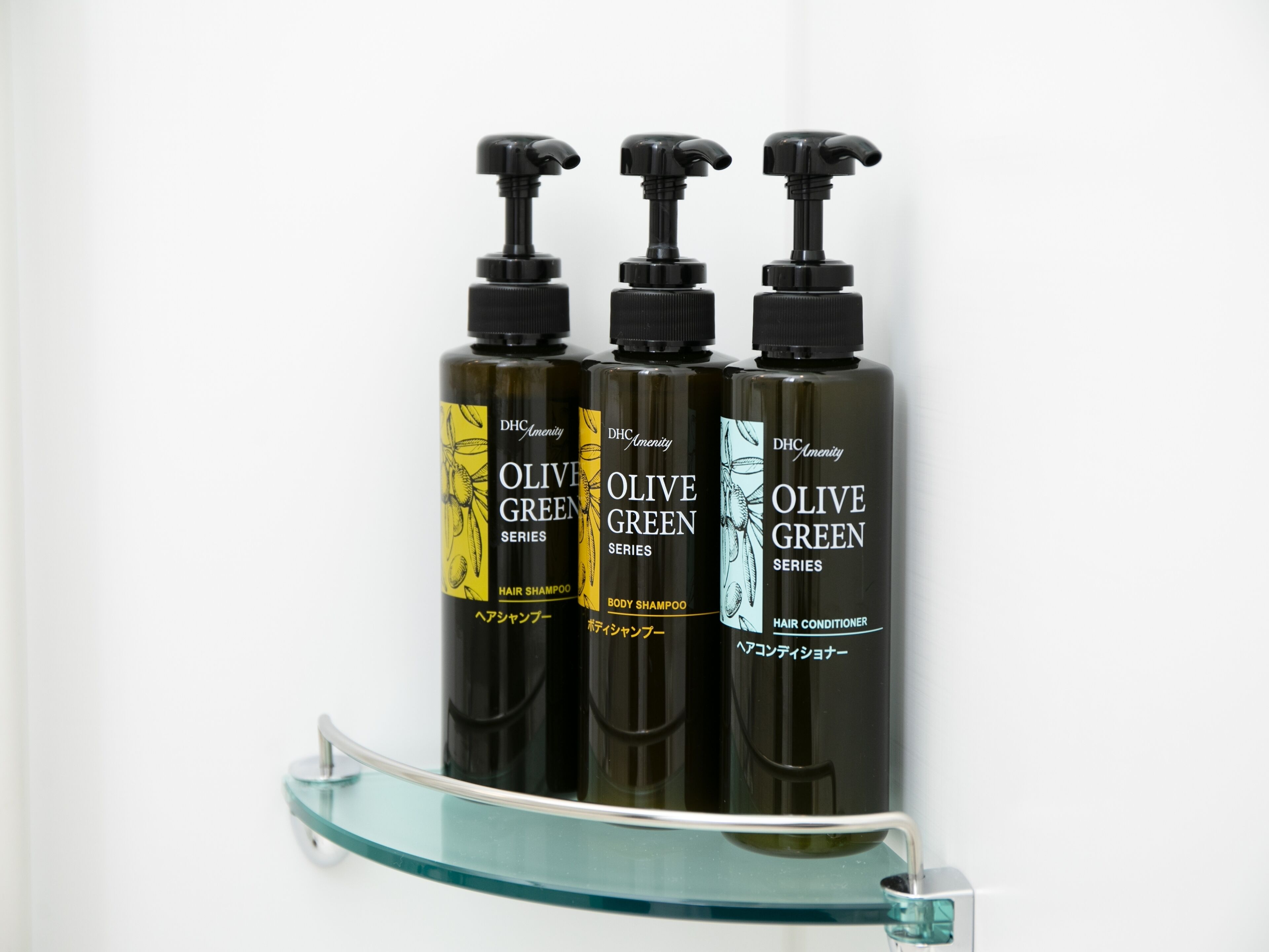 Tre flaconi di prodotti per capelli DH Amenity serie Verde Oliva su un ripiano in vetro.